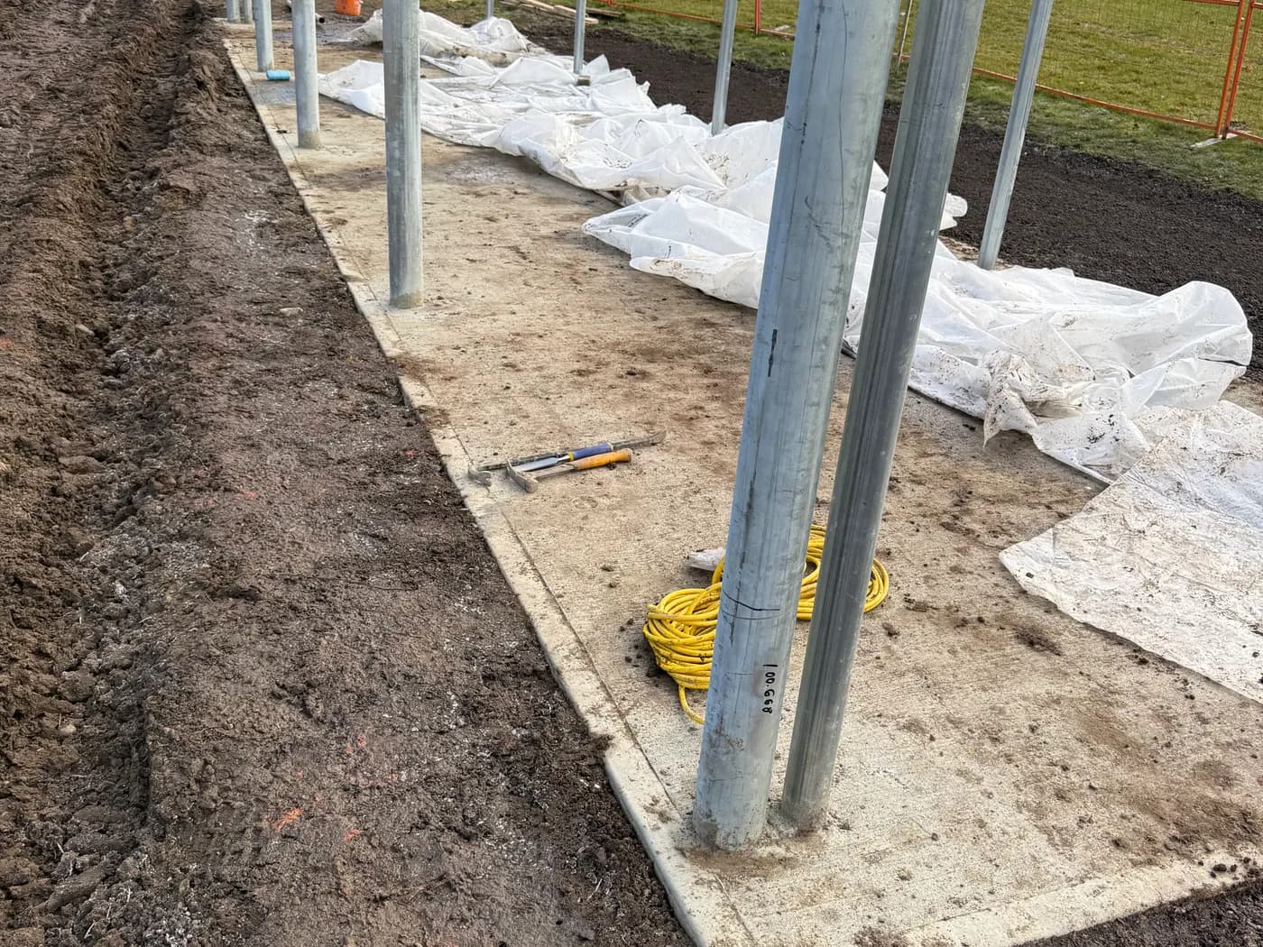 Newly poured concrete footing with galvanized posts set to line and grade, trench backfill alongside