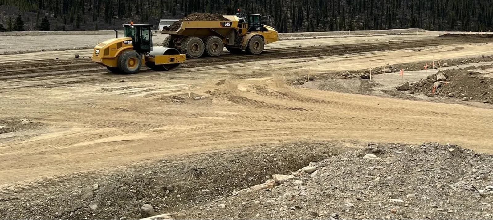 Smooth drum packer compacting a large graded building pad while an articulated rock truck hauls structural fill, with grade stakes set