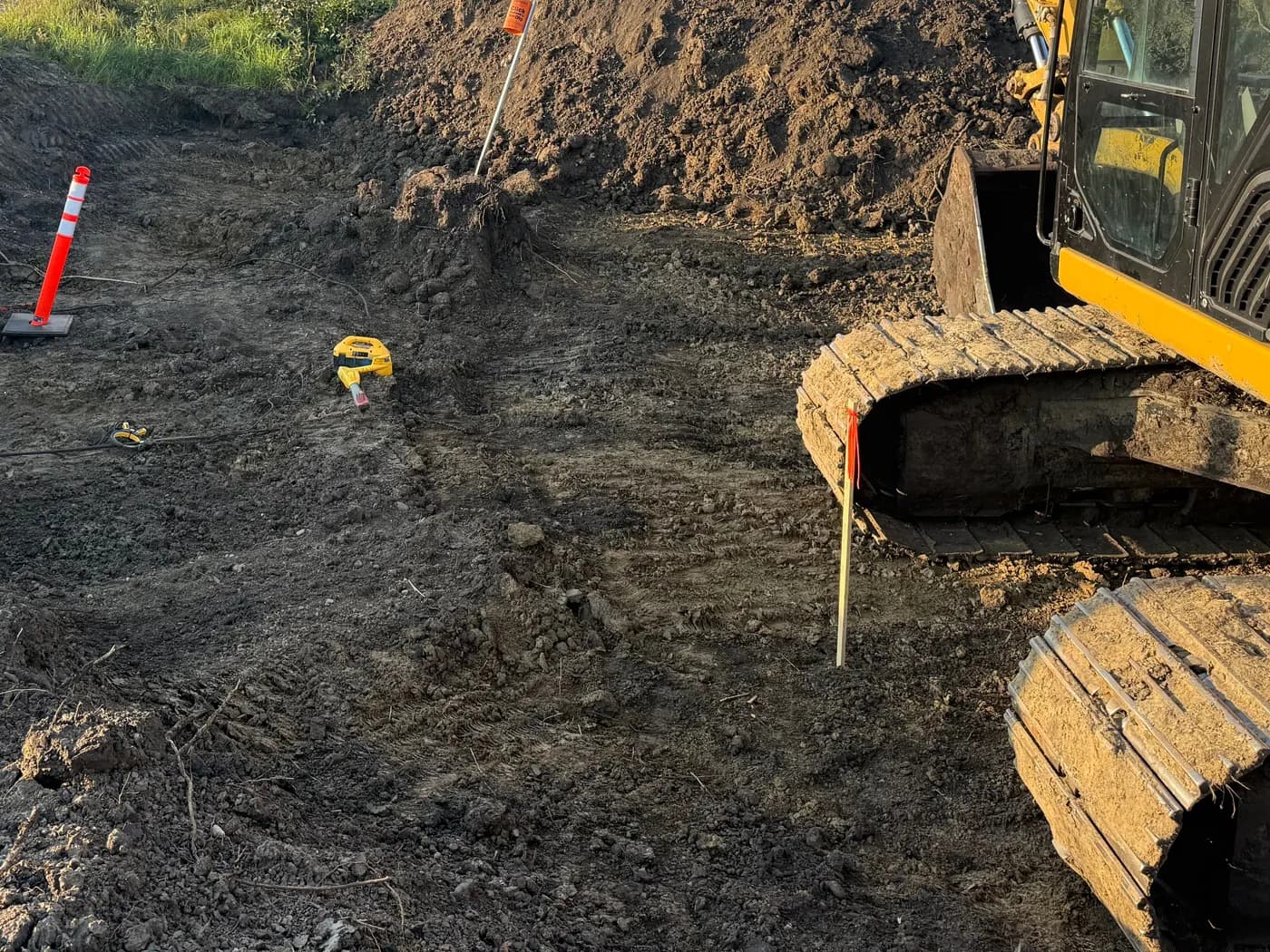 Excavator trenching through dark organic soil with layout stakes and marking whiskers set
